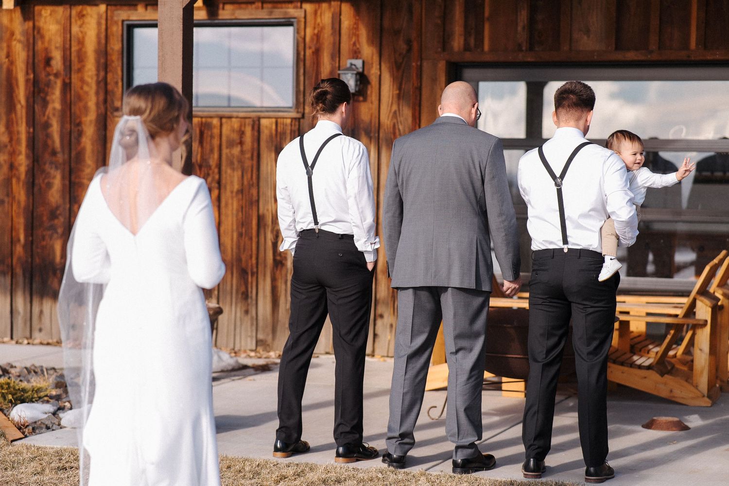 Wedding party in formal attire stands near rustic wooden building with groomsmen wearing black suspenders.