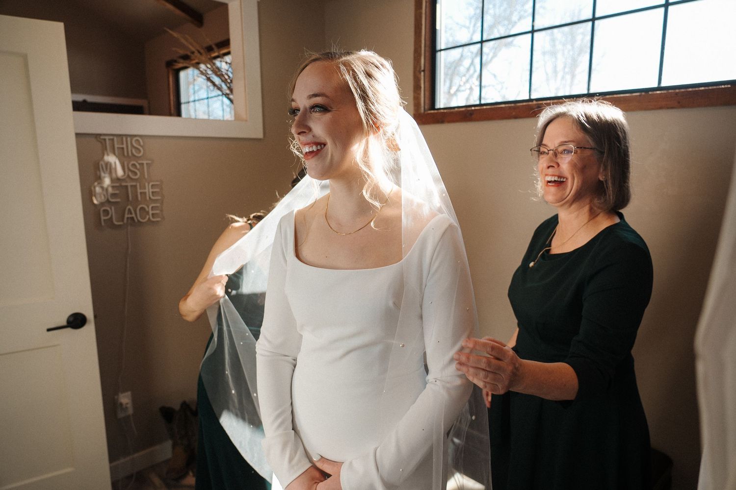 A joyful bride wearing a long-sleeved white wedding dress laughs while getting ready in a sunlit room.