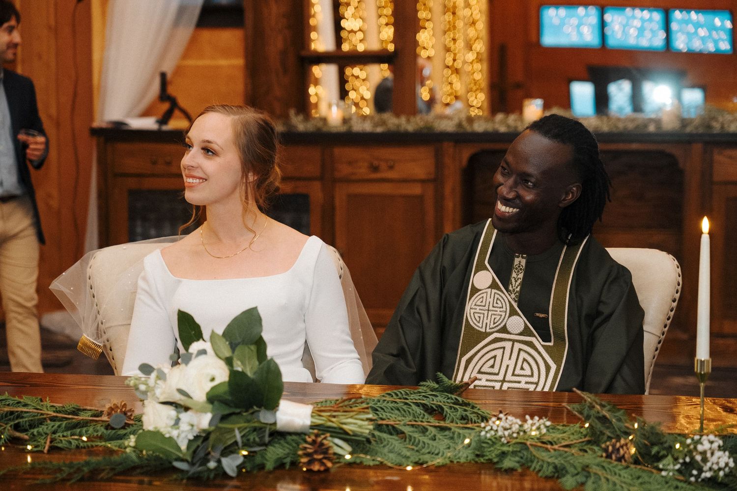 Happy newlyweds smile and laugh together at their reception table decorated with greenery and white flowers.