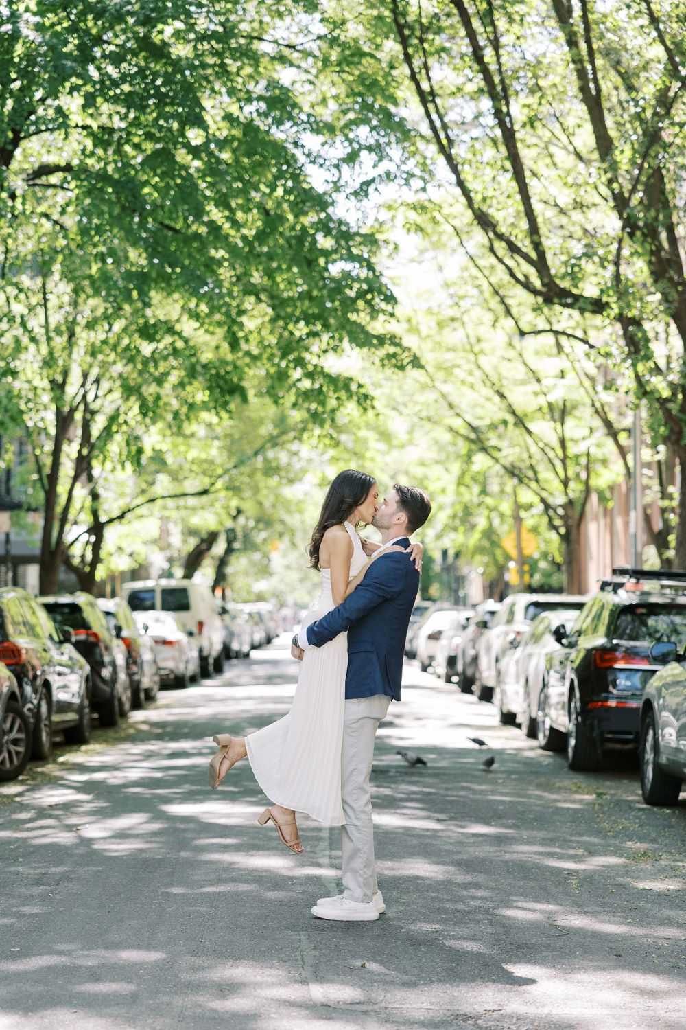 A romantic street scene with couple embracing under tree-lined avenue with parked cars in New York City.