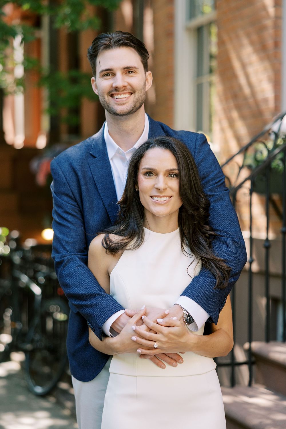 Engaged couple in formal attire pose together against brick building in natural light in New York City.
