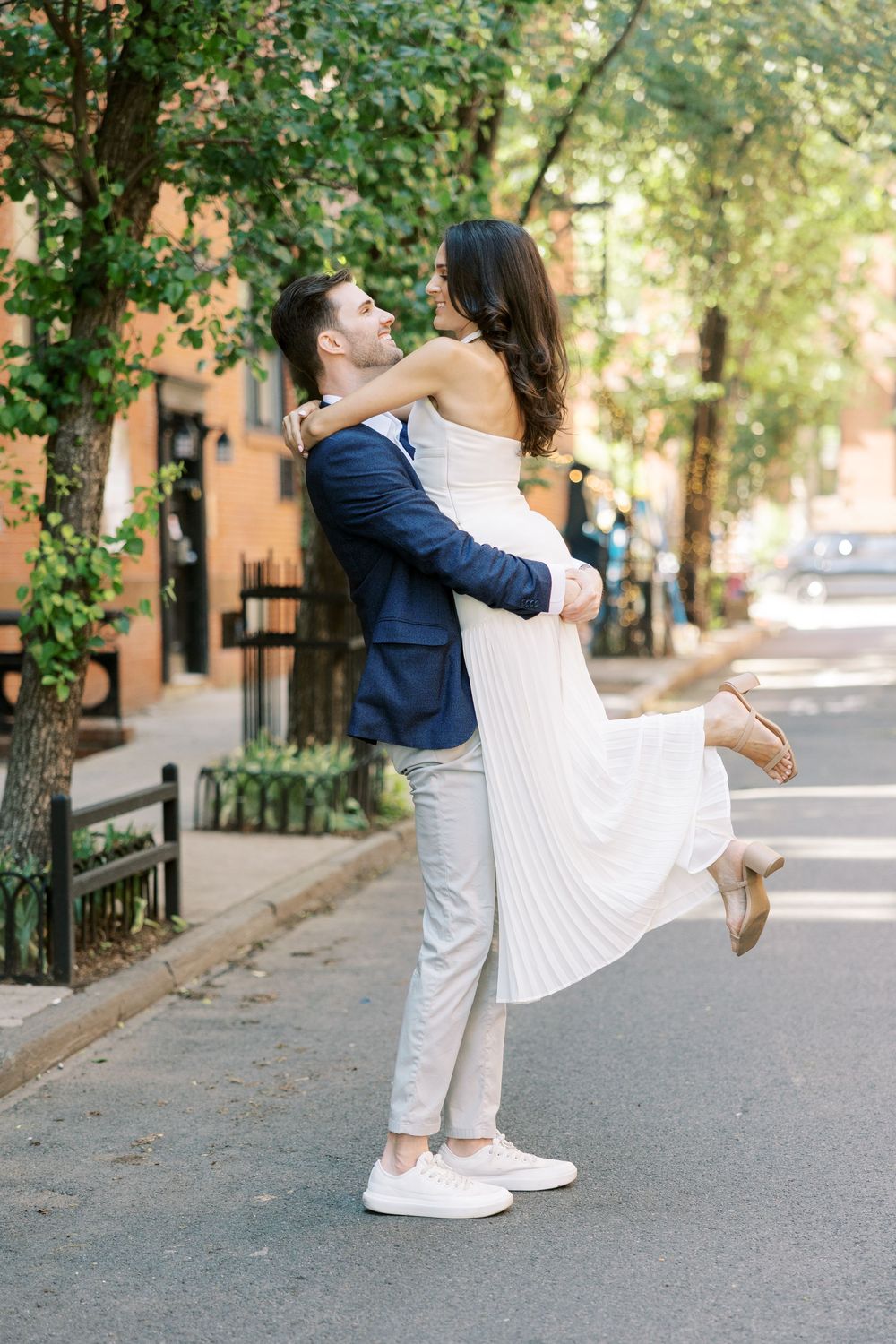Couple in formal wear share playful lift moment on tree-lined city street in New York City.
