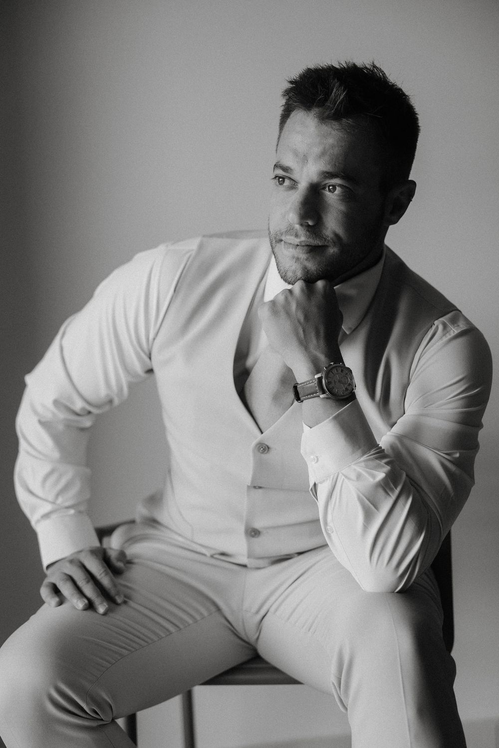 Professional black and white fashion portrait of a man in white dress shirt and vest sitting on a chair.