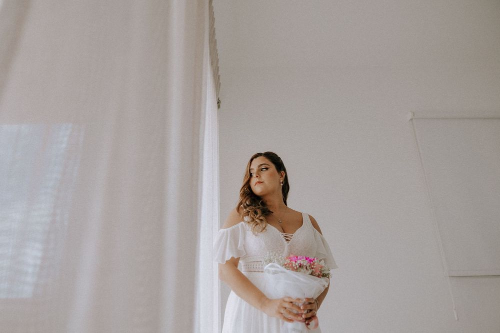 Woman in a flowing white dress stands by a window in a bright minimalist room.