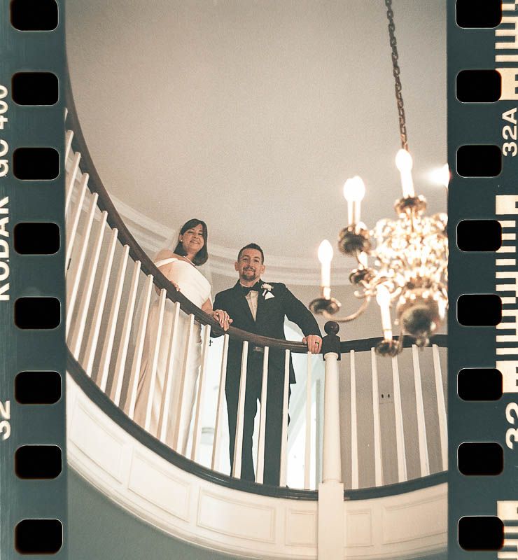 Elegant Wedding couple poses on a curved staircase with white railings under a crystal chandelier Kodak Gold 400 Film Pittsburgh PA.