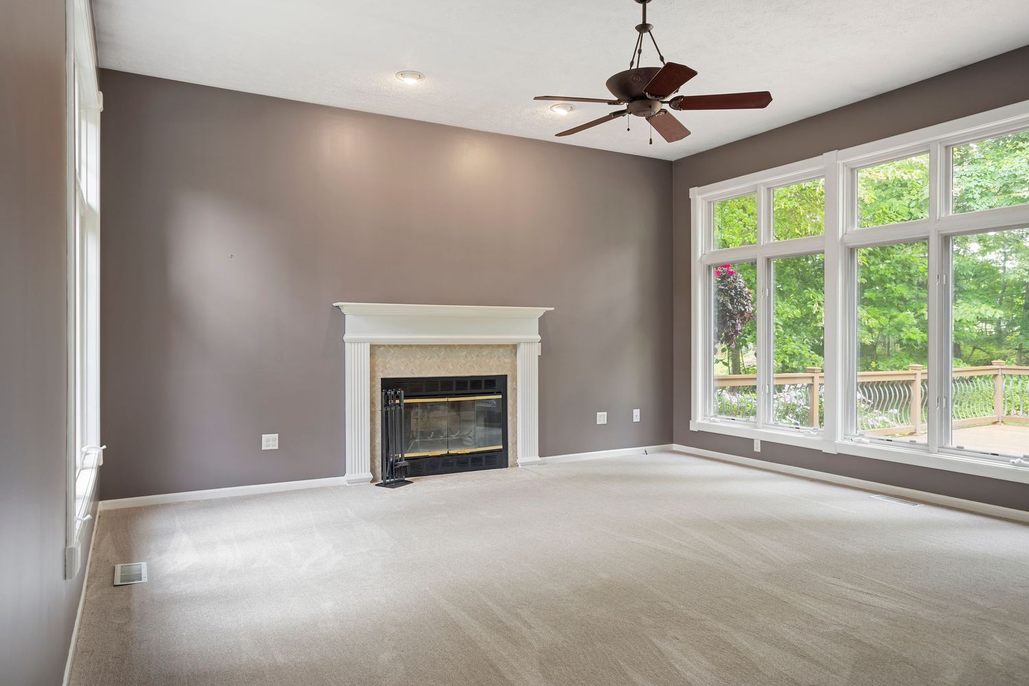 Large living room with gray walls, white fireplace mantel, ceiling fan, and wall of windows.