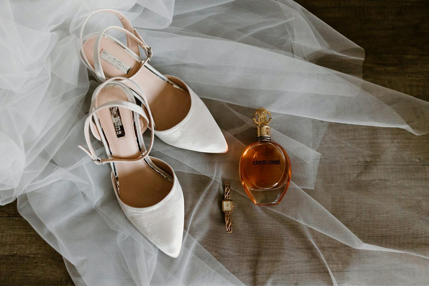 White bridal heels and perfume bottle arranged on delicate tulle veil against dark background.