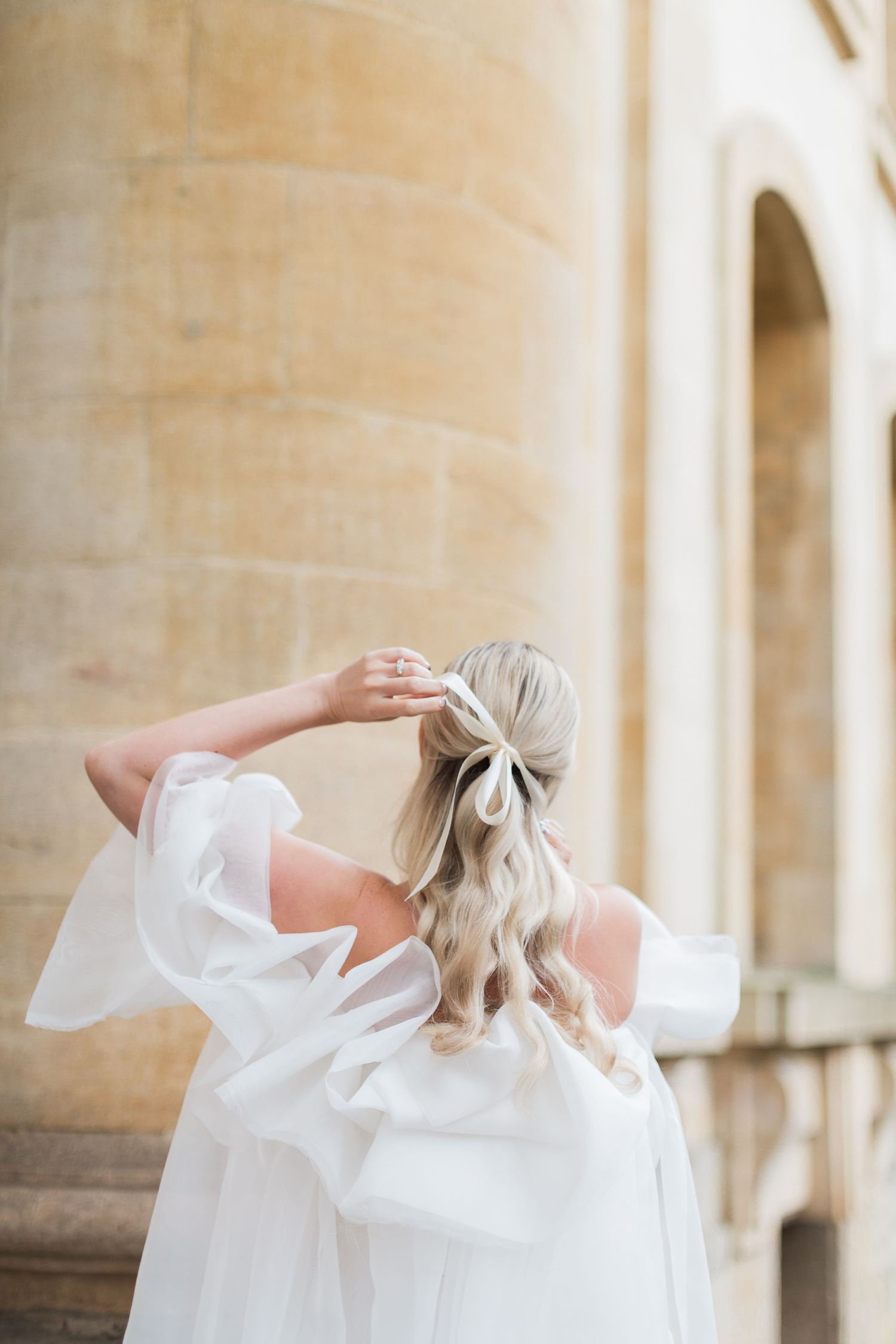 Back view of bride in flowing white dress with long blonde hair and bow detail standing against stone architecture Oxford elopement