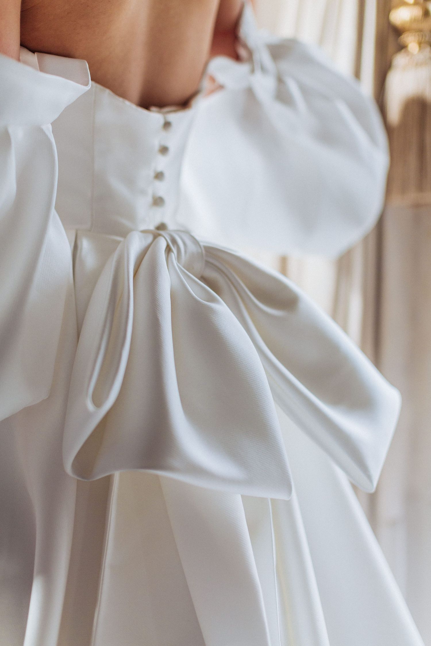 Close-up view of an elegant white wedding dress detail featuring an oversized bow and button closure by Hayley Elizabeth. Hedsor House