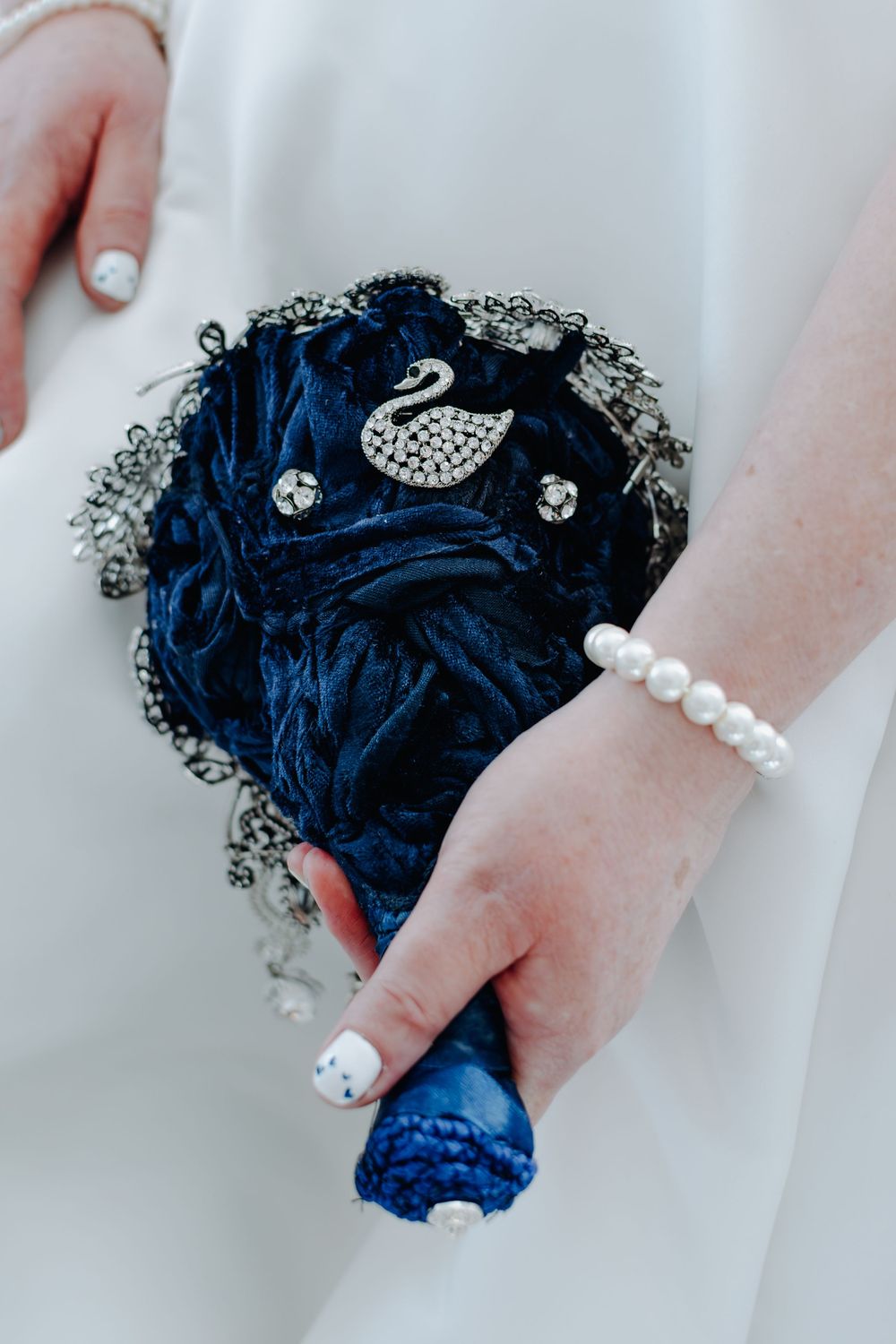 Navy blue satin bouquet with silver swan brooch, black lace trim and pearl bracelet detail against white fabric.