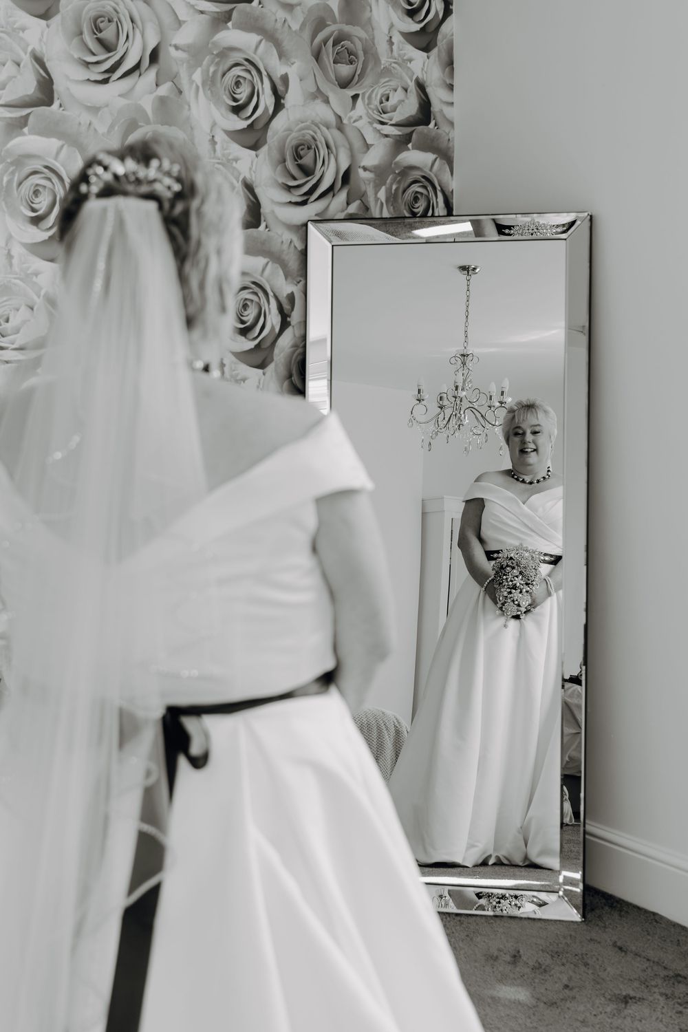 Elegant wedding dress reflection in full-length mirror with floral wallpaper backdrop and chandelier in black and white.