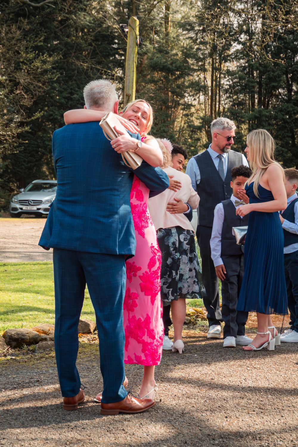 Wedding guests in formal attire gather on a gravel path surrounded by trees during an outdoor celebration.