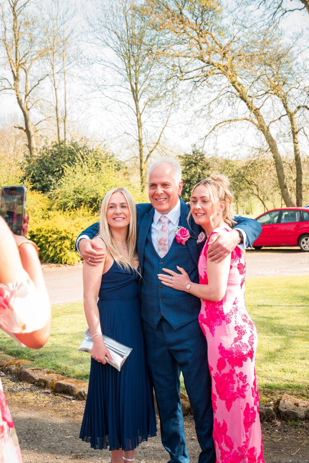 Three people pose together outdoors at a formal event on a sunny day with trees and a parked car in the background.