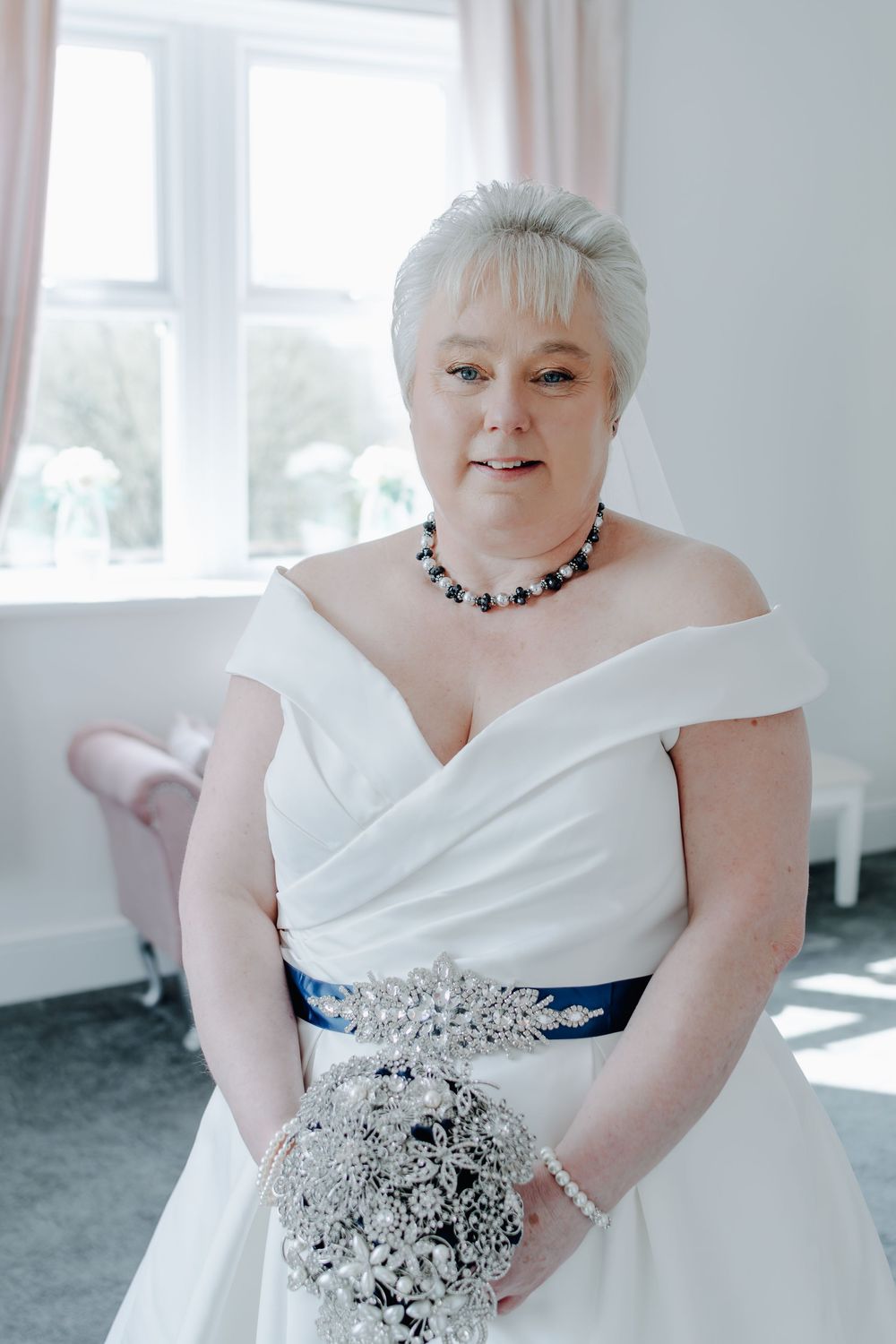 A bride in an elegant off-shoulder white wedding gown with a navy sash and crystal brooch holds a silver bouquet.