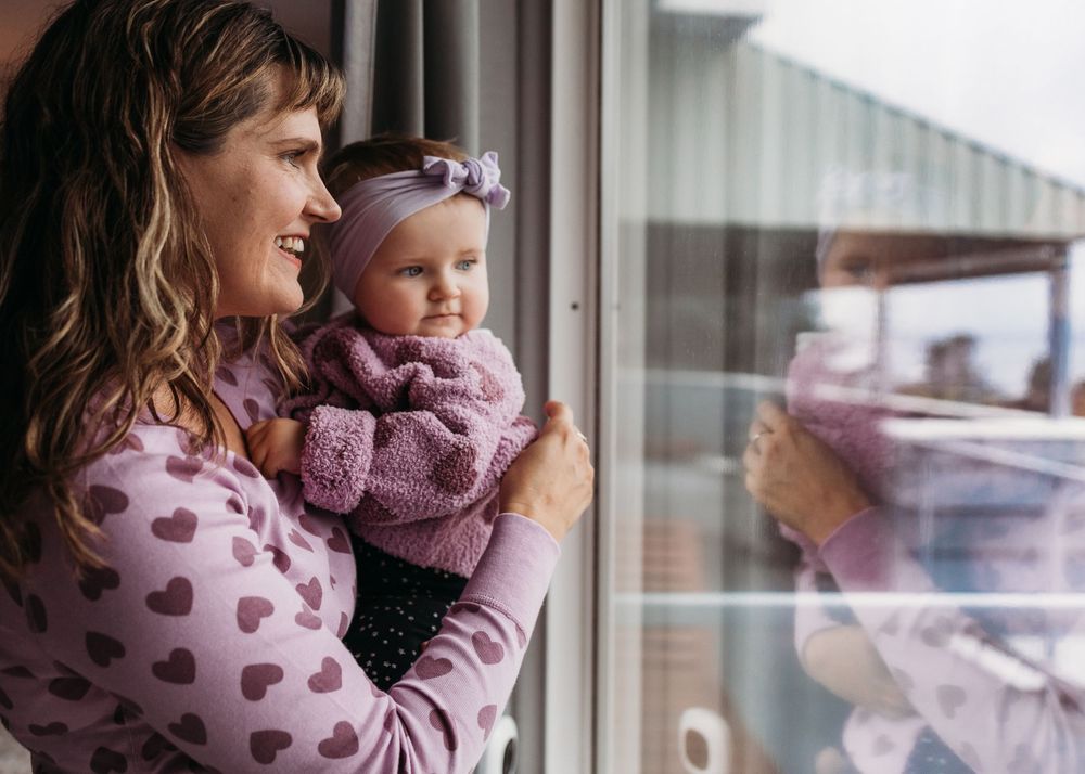 A parent and baby wearing purple sweaters look out a window together on a cloudy day.