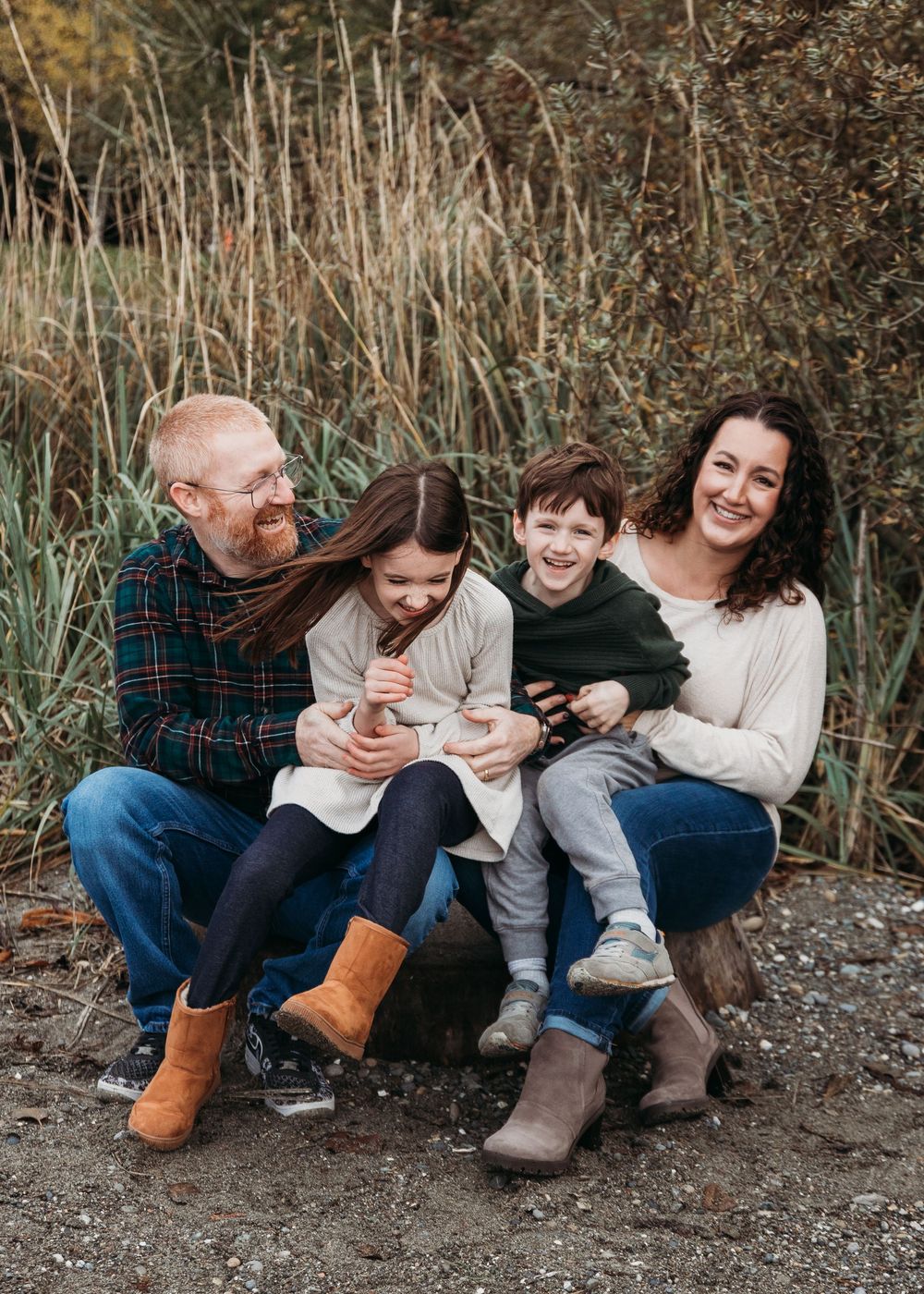 A family laughs together while sitting on rocks near tall grass and reeds during an outdoor autumn photo session.