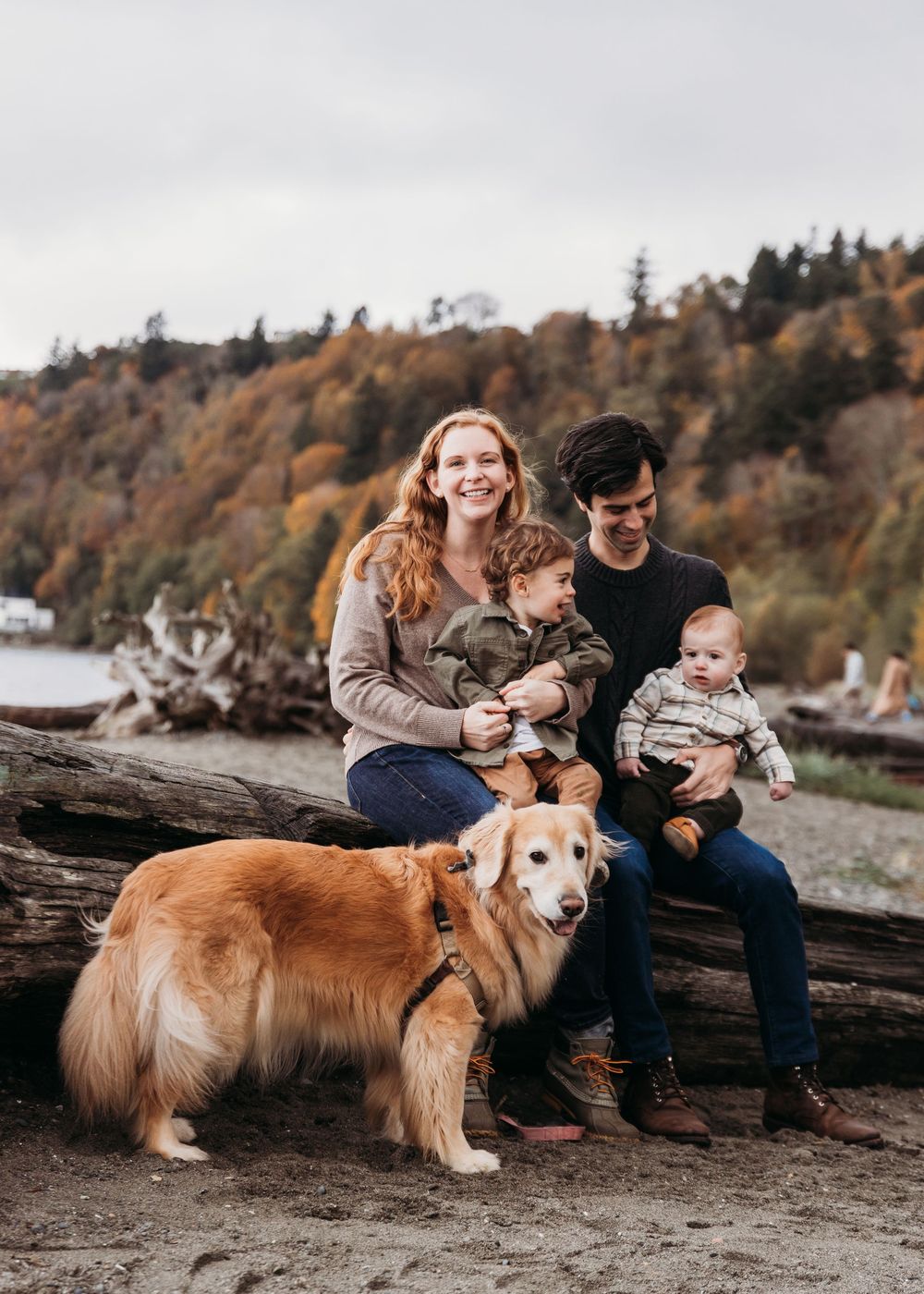 Family sitting on driftwood at beach with golden retriever dog during autumn with fall foliage in background.