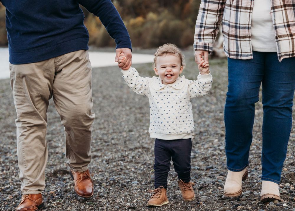 Happy toddler in a polka dot sweater stands outdoors holding hands with people on a gravel path.