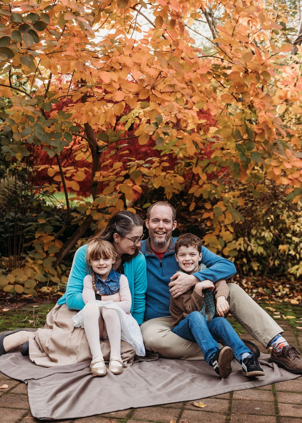 A group sits together on a blanket against vibrant orange and red autumn foliage in a backyard setting.