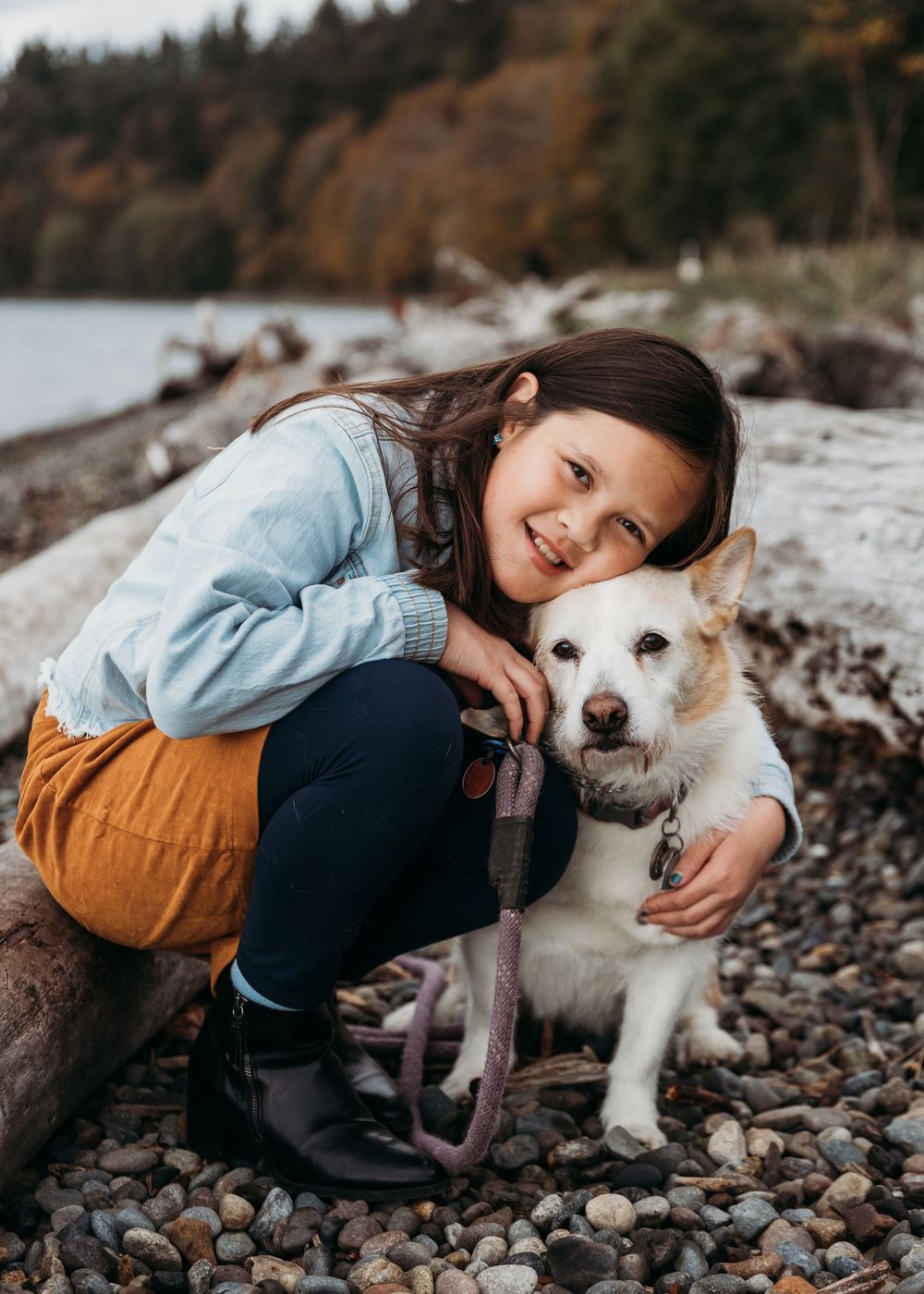 A young child and white dog share a sweet moment together on a rocky beach shore with autumn trees in the background.