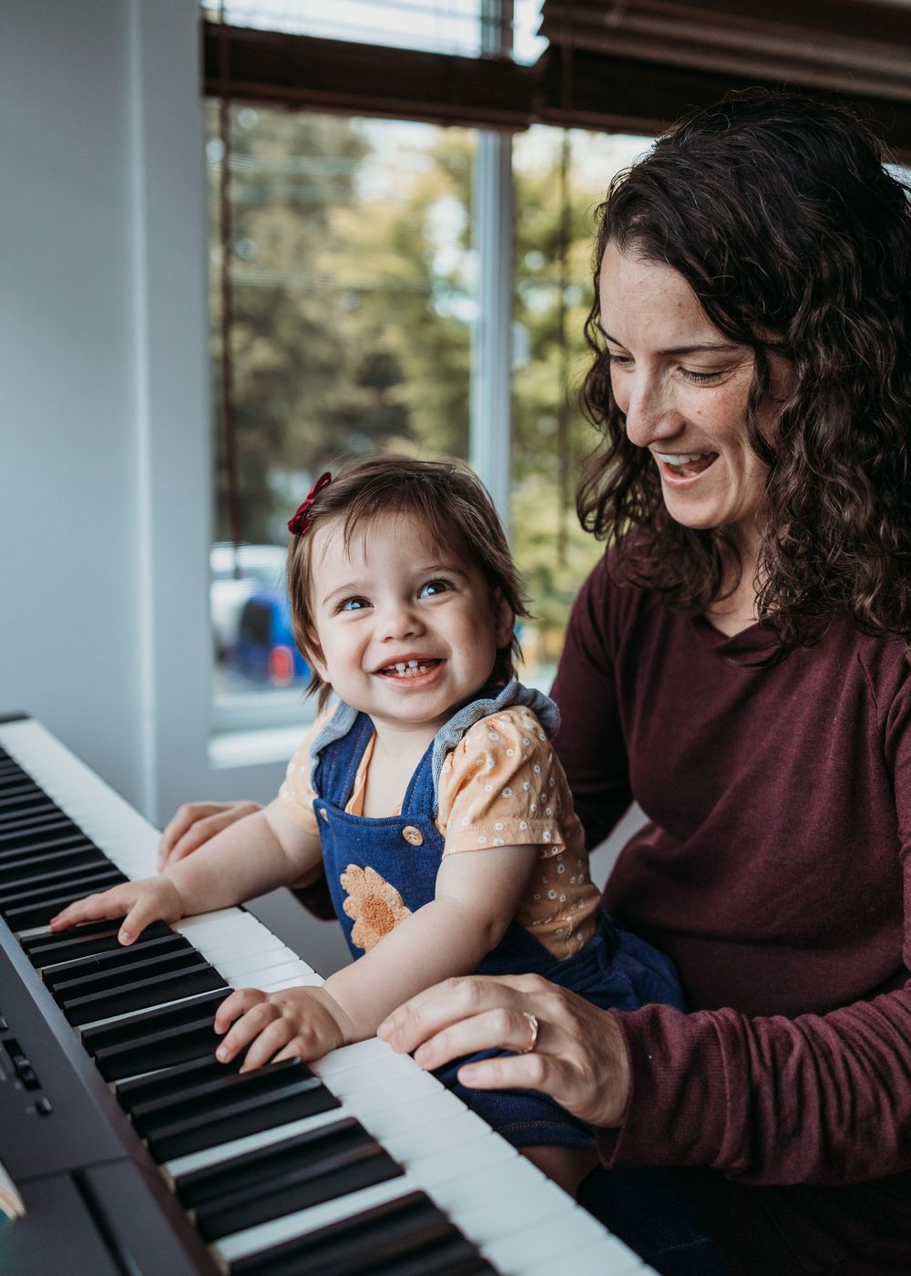 Adult and young child sit together playing an electronic keyboard and smiling by a window.