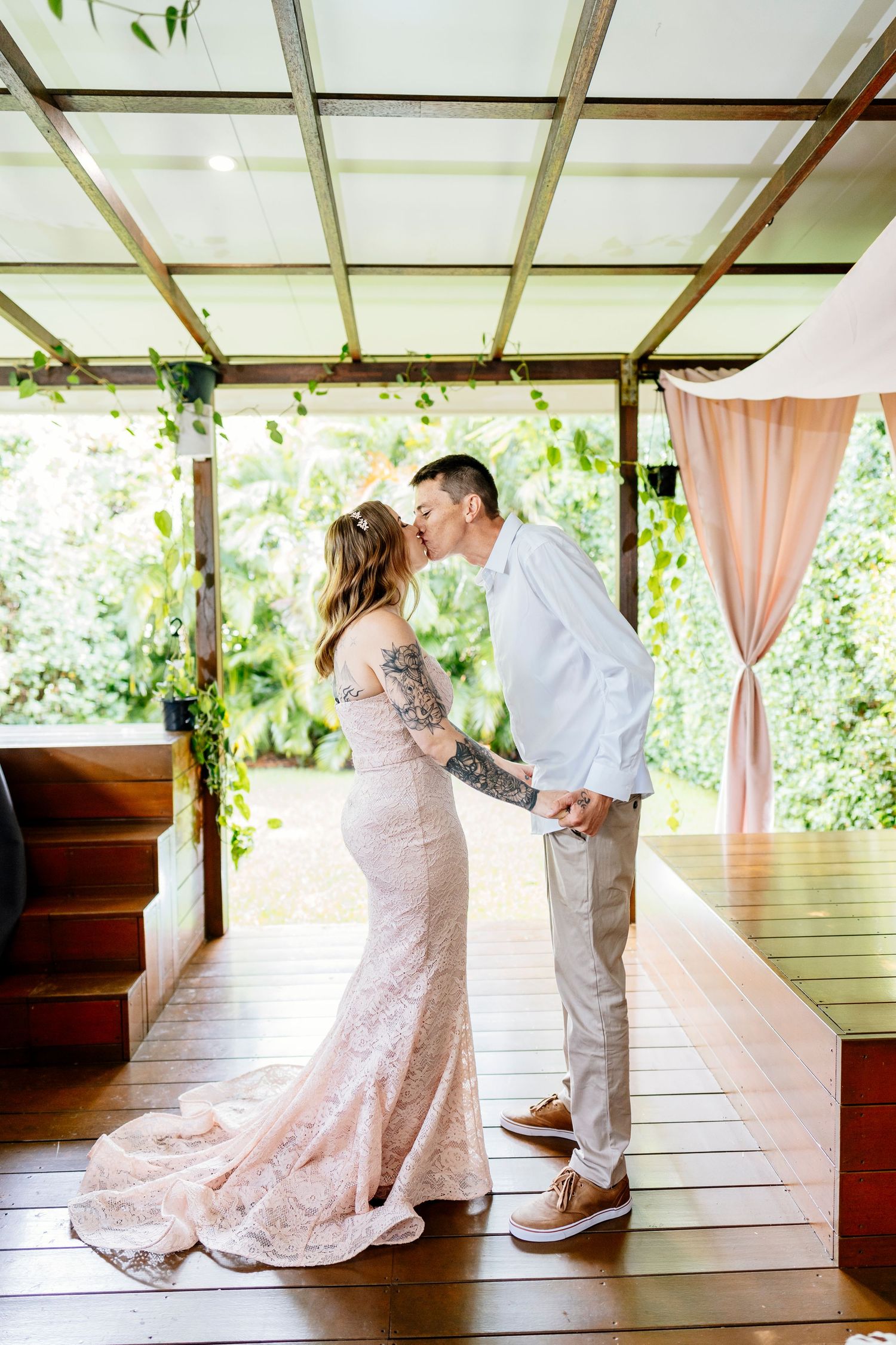 Newlyweds share a romantic kiss on wooden bridge by willow tree with white roses bouquet and elegant wedding attire.