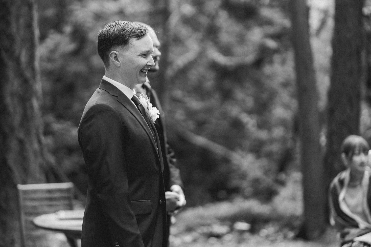 Joyful groom laughs during an outdoor wedding ceremony in a wooded setting captured in black and white.