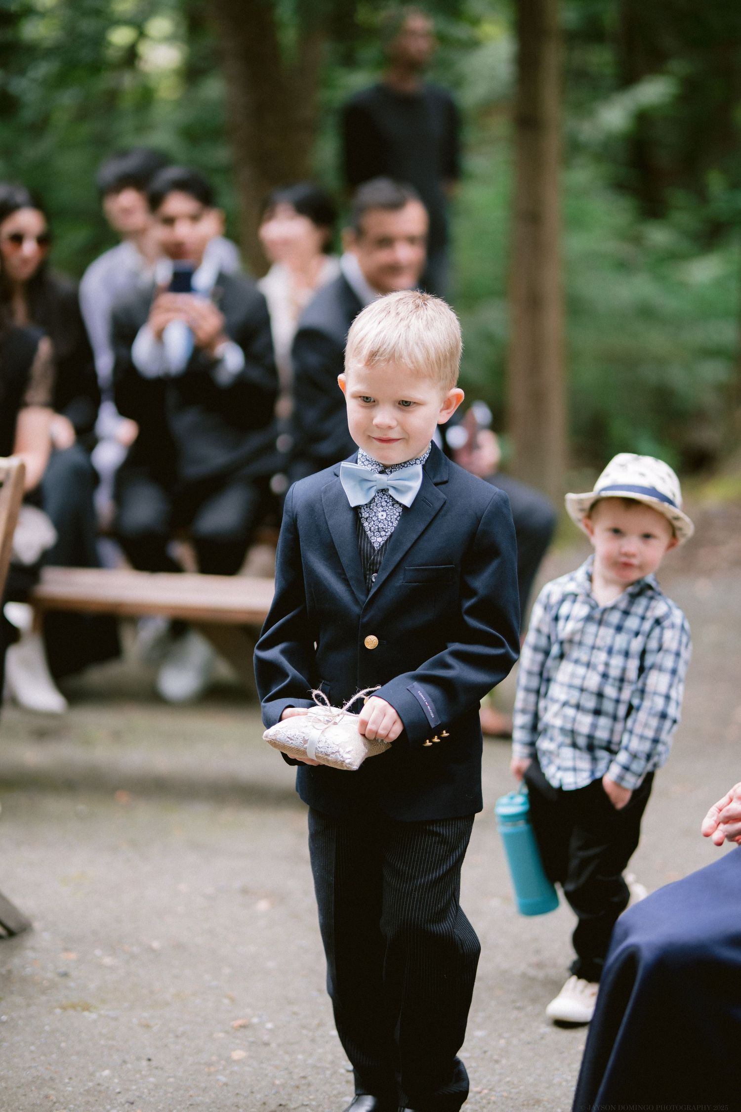Young child in formal navy suit with bowtie walking down outdoor path at wedding ceremony.