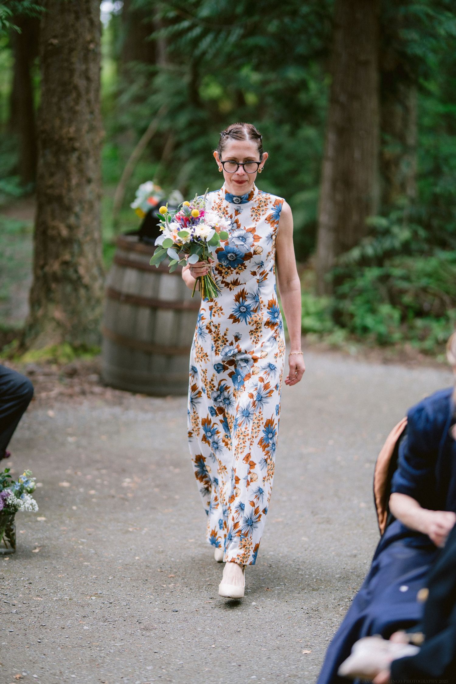A person in a floral print dress walks down a forest path carrying a colorful bouquet of flowers.