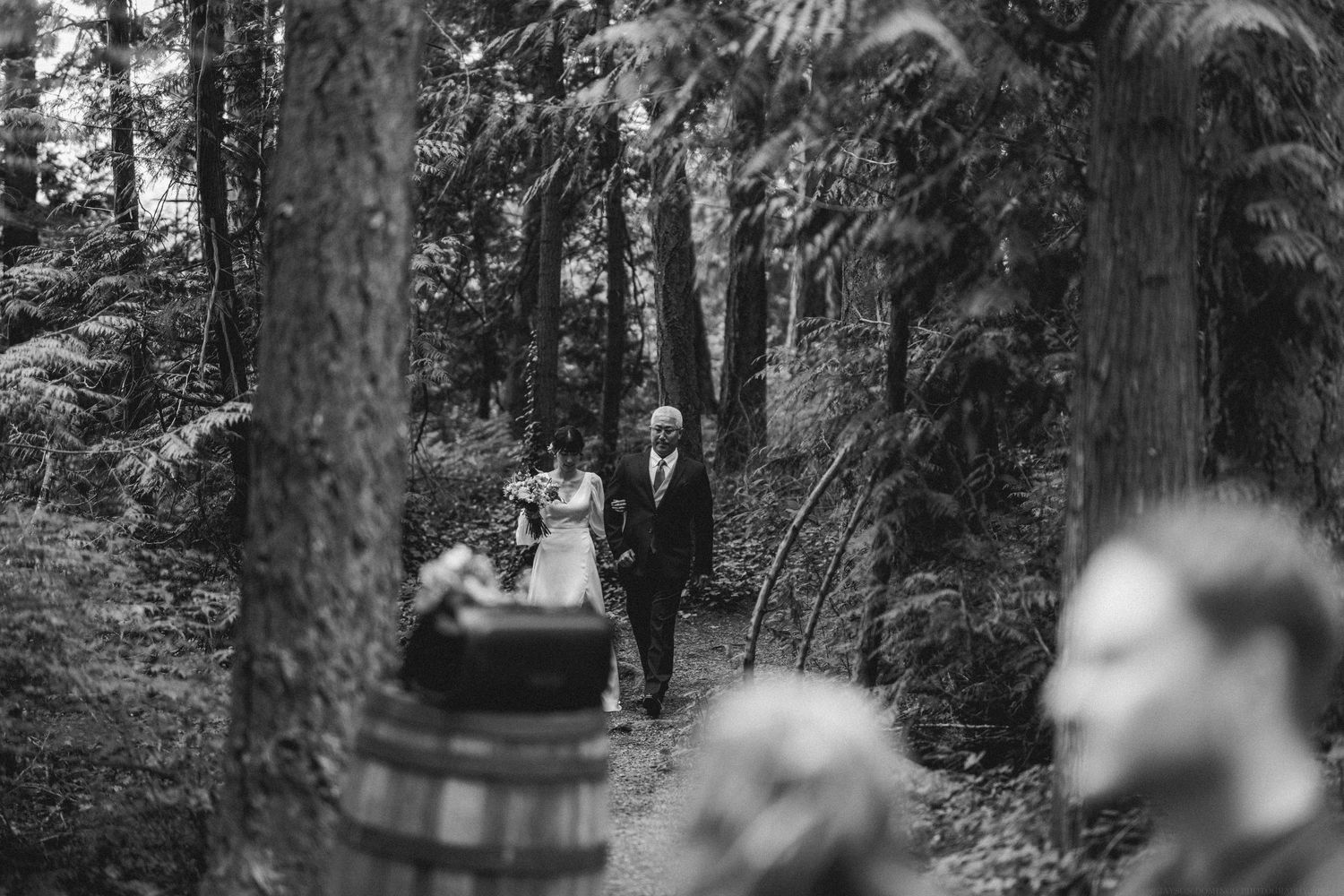 Black and white photograph of a forest wedding ceremony path with guests walking among tall trees.