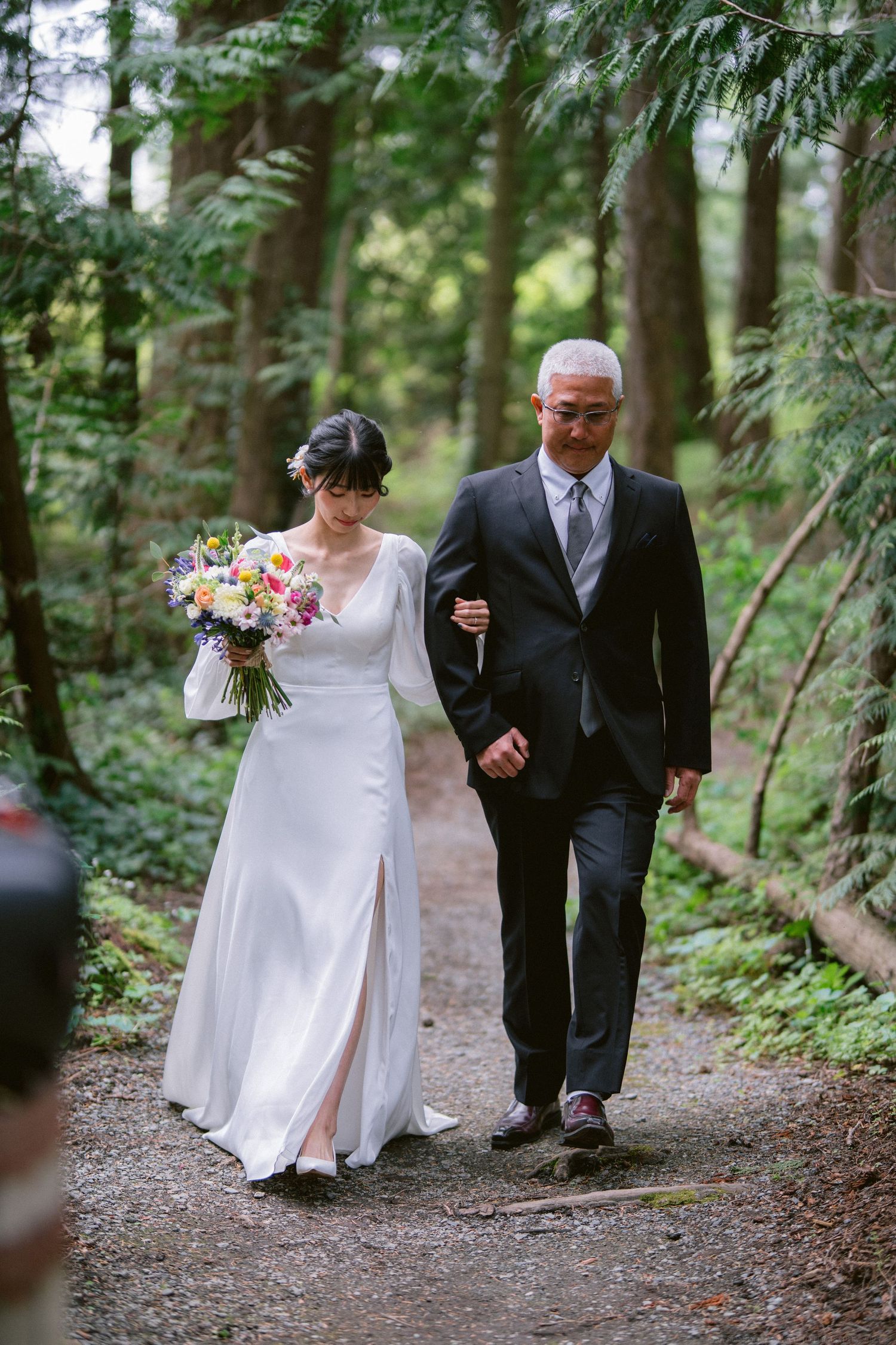 A couple walks arm in arm down a wooded path, with the bride in white carrying a floral bouquet.