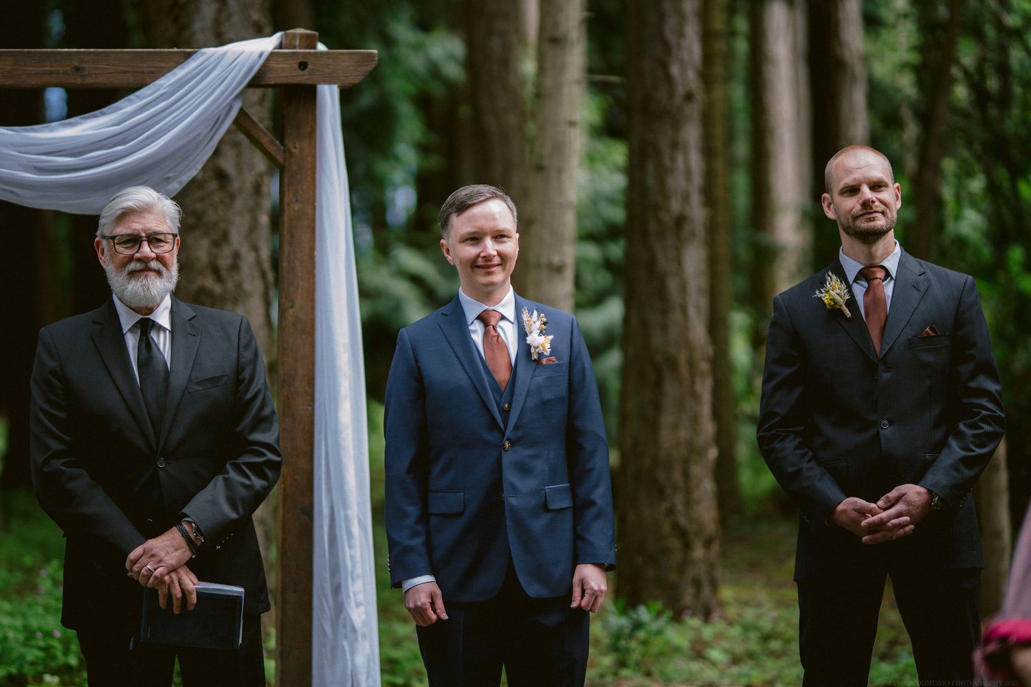 Three men in formal suits stand at an outdoor wedding ceremony site in a lush forest setting.