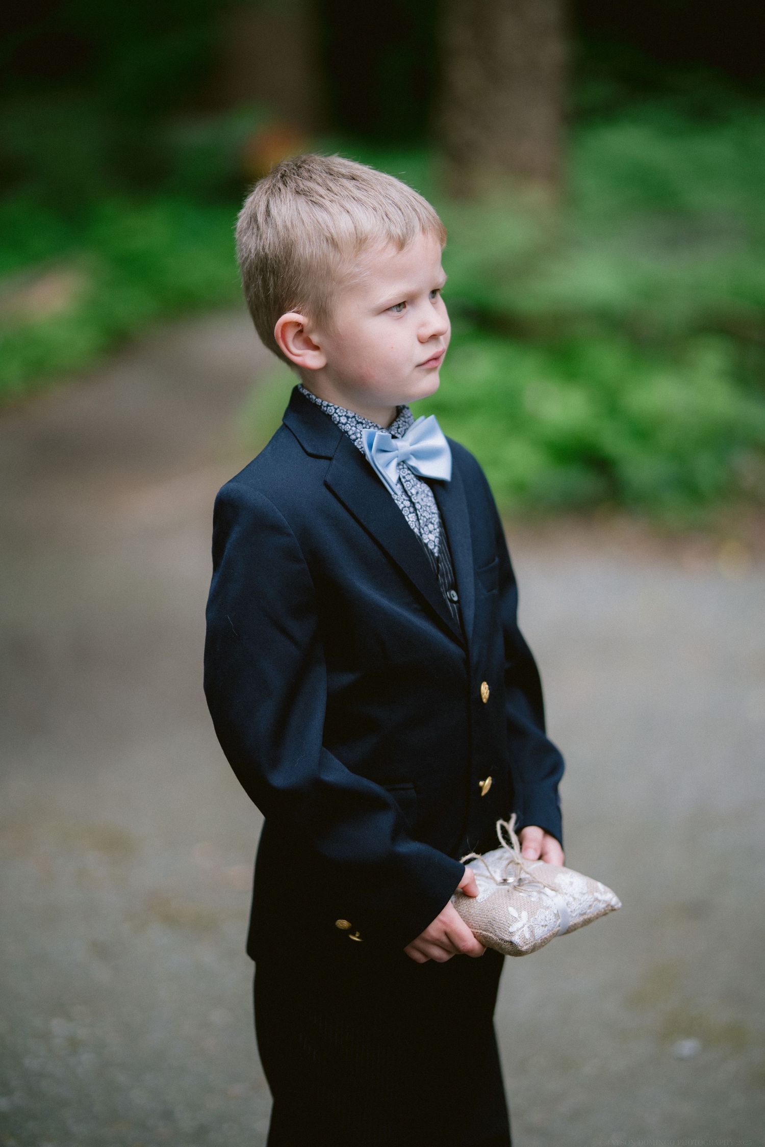 A young ring bearer in a navy suit holds a small pillow while standing outdoors in a wooded setting.