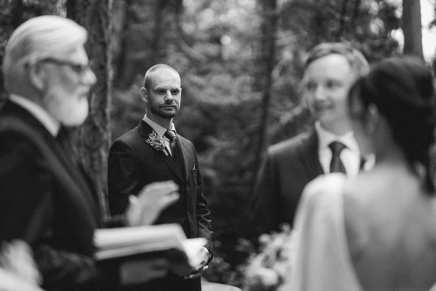 A black and white candid moment during an outdoor wedding ceremony with people standing in a wooded setting.