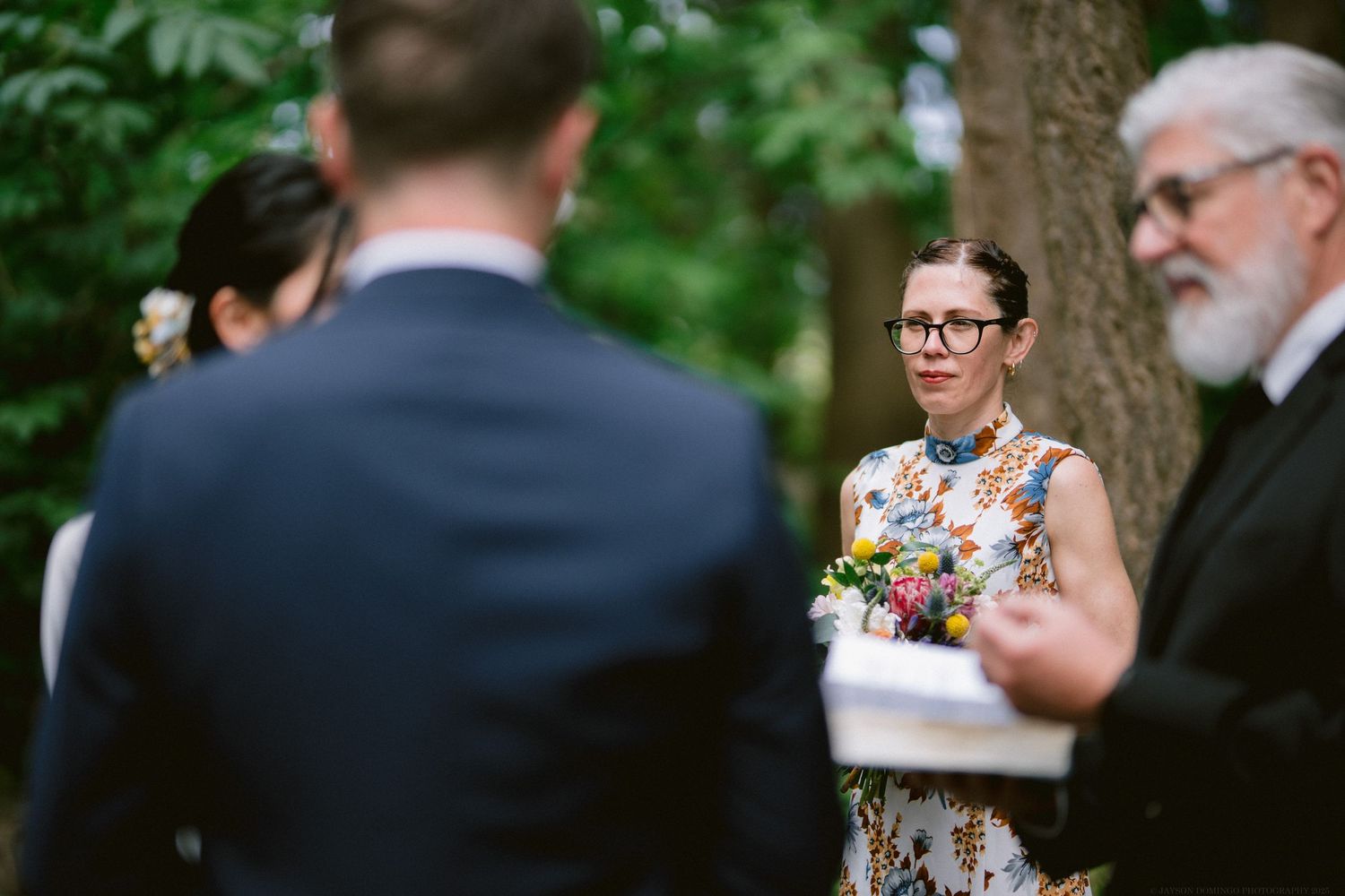 Wedding ceremony outdoors with minister and colorful floral dress against green foliage background.