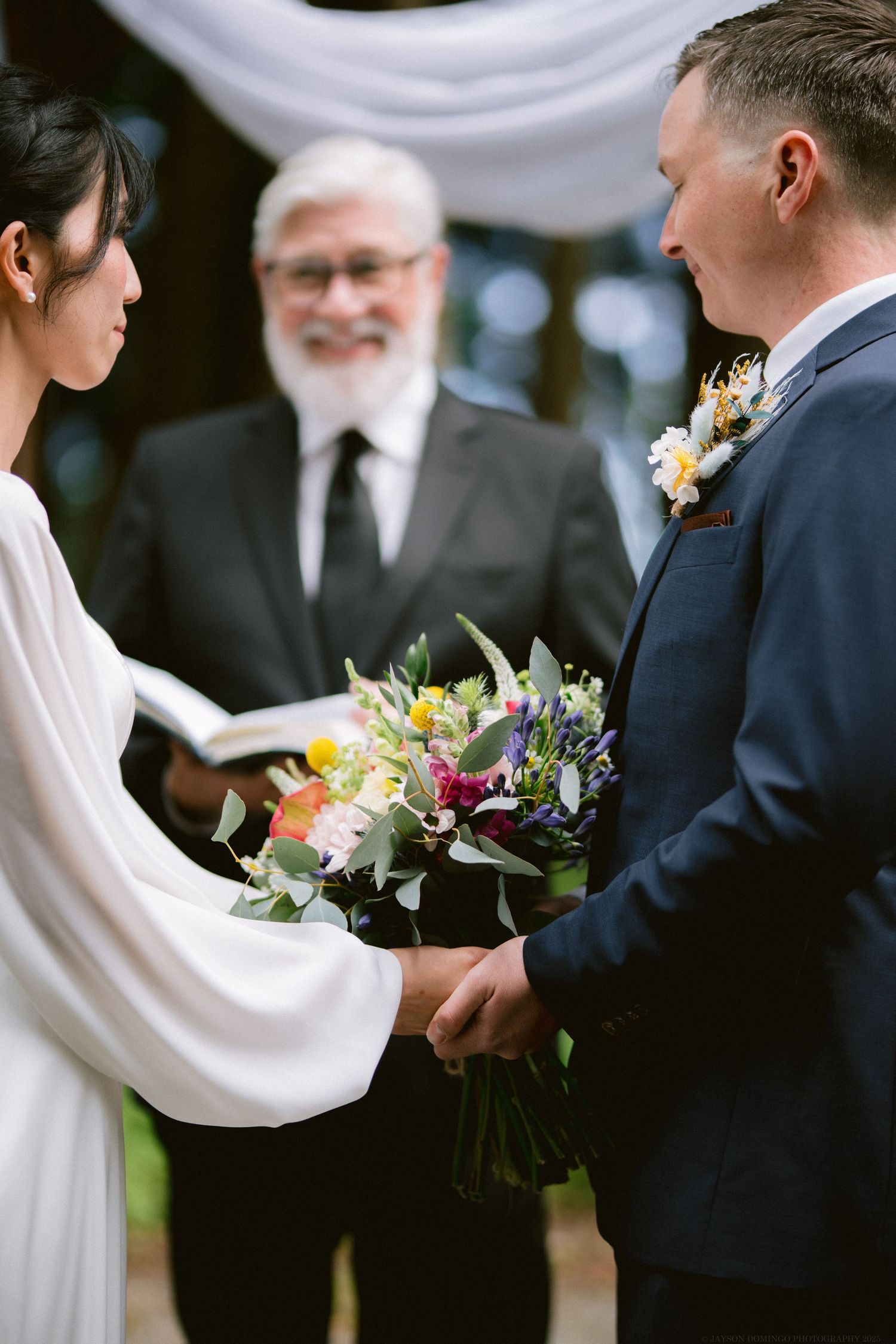 A couple exchanges wedding vows in front of an officiant while the bride holds a vibrant mixed floral bouquet.