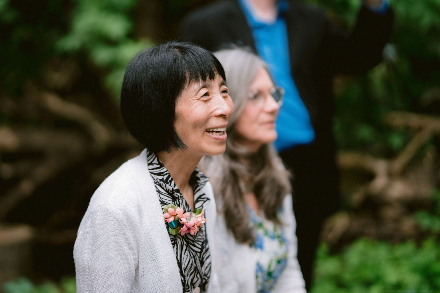 Someone laughs joyfully while wearing a floral corsage at an outdoor event with greenery in the background.