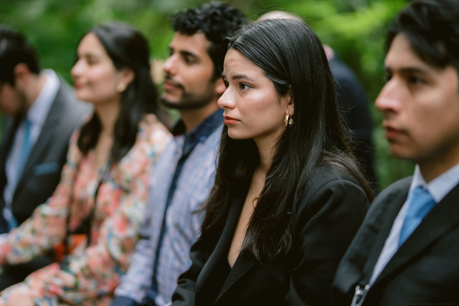 Wedding guests in formal attire sit solemnly during an outdoor ceremony.