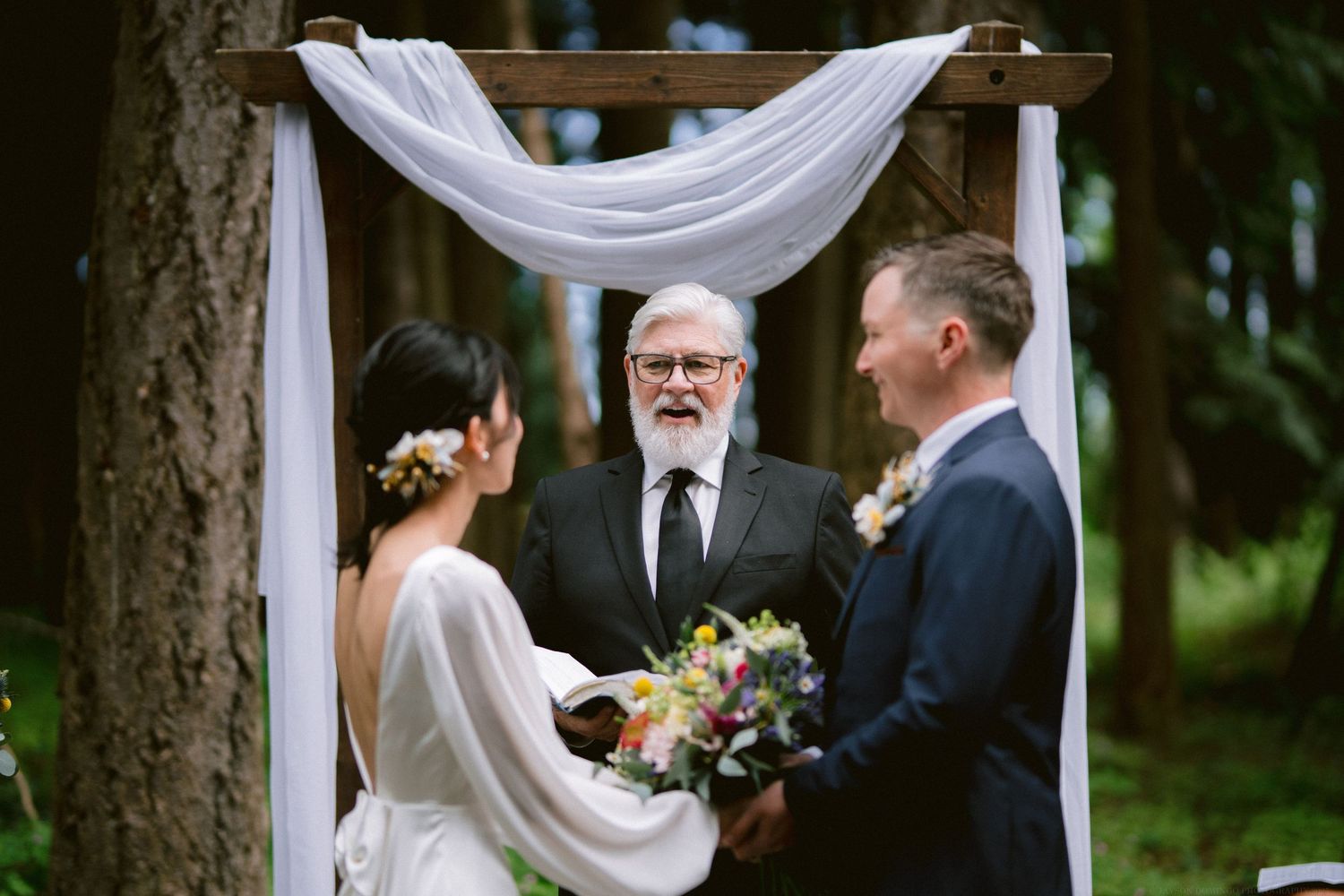 A couple exchanges vows under a white draped arch during an intimate forest wedding ceremony.