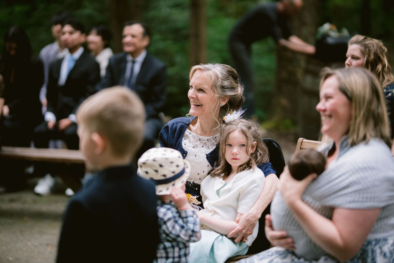 Wedding guests react with joy and laughter during an outdoor ceremony in a wooded setting.