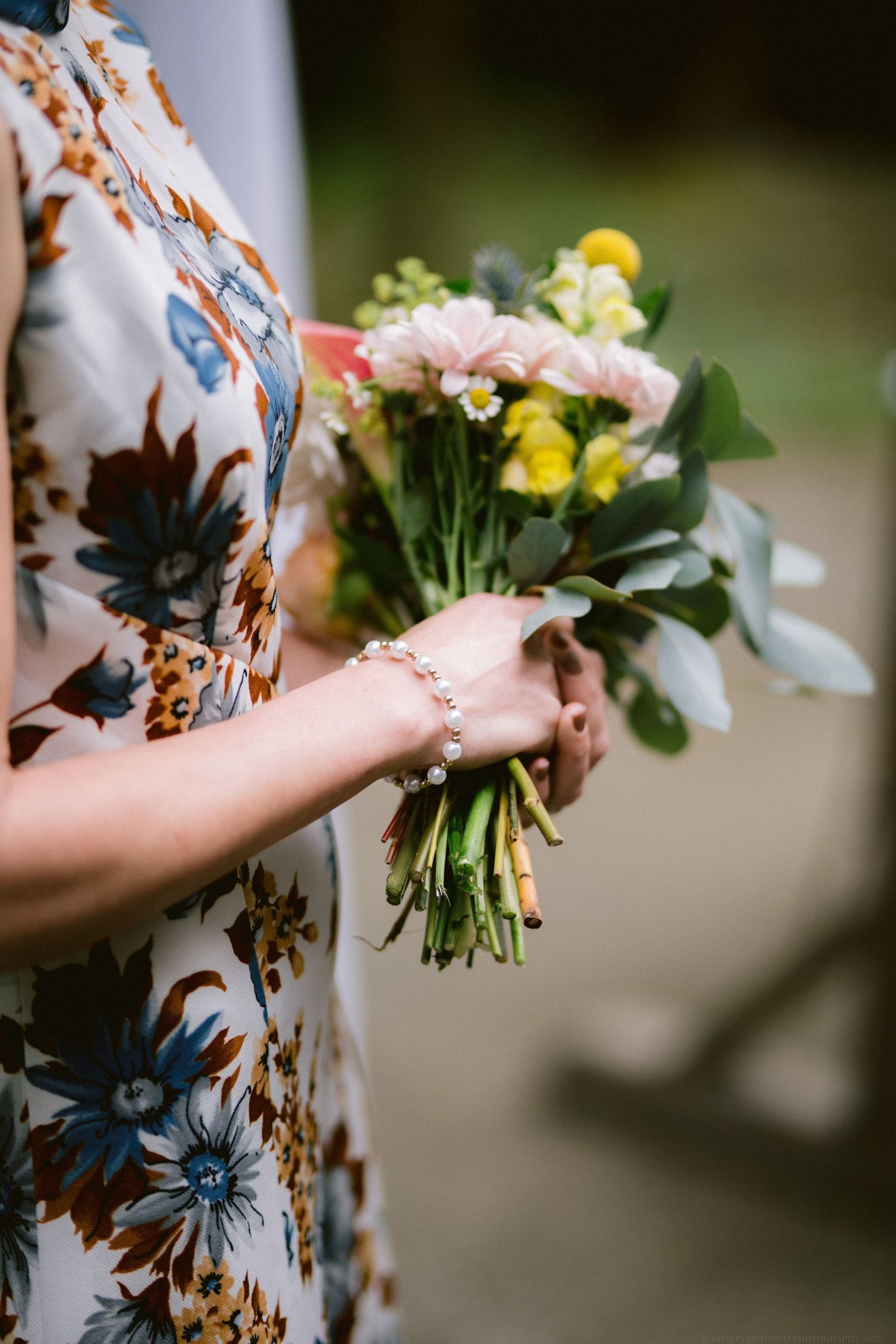 A person wearing a floral dress holds a delicate bouquet of pink and yellow flowers with greenery.