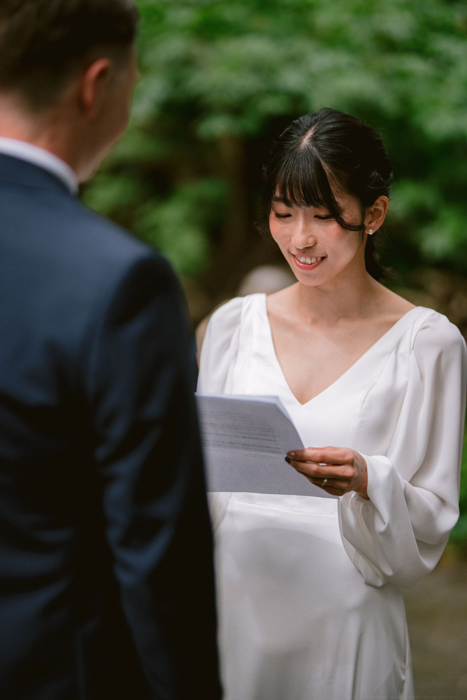A bride in a white dress reads from paper during an outdoor forest wedding ceremony.