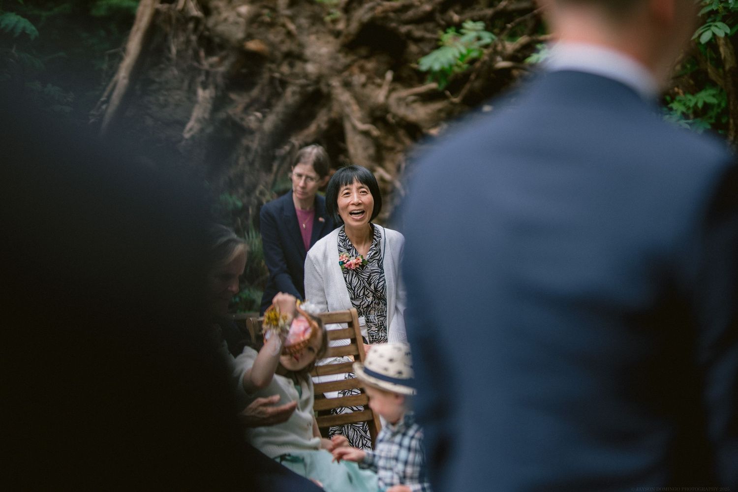 Wedding guests seated outdoors among trees share a light moment during an intimate forest ceremony.