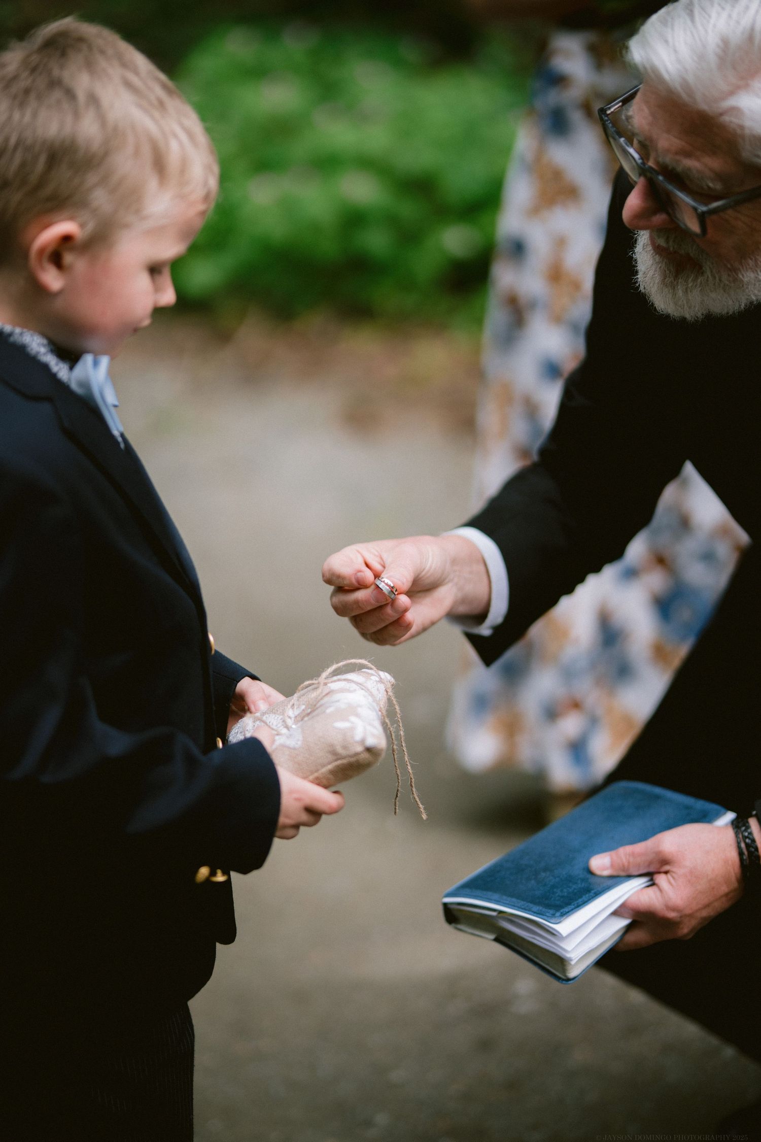 An older person in a dark suit hands something delicate to a young child in formal attire while holding a book.