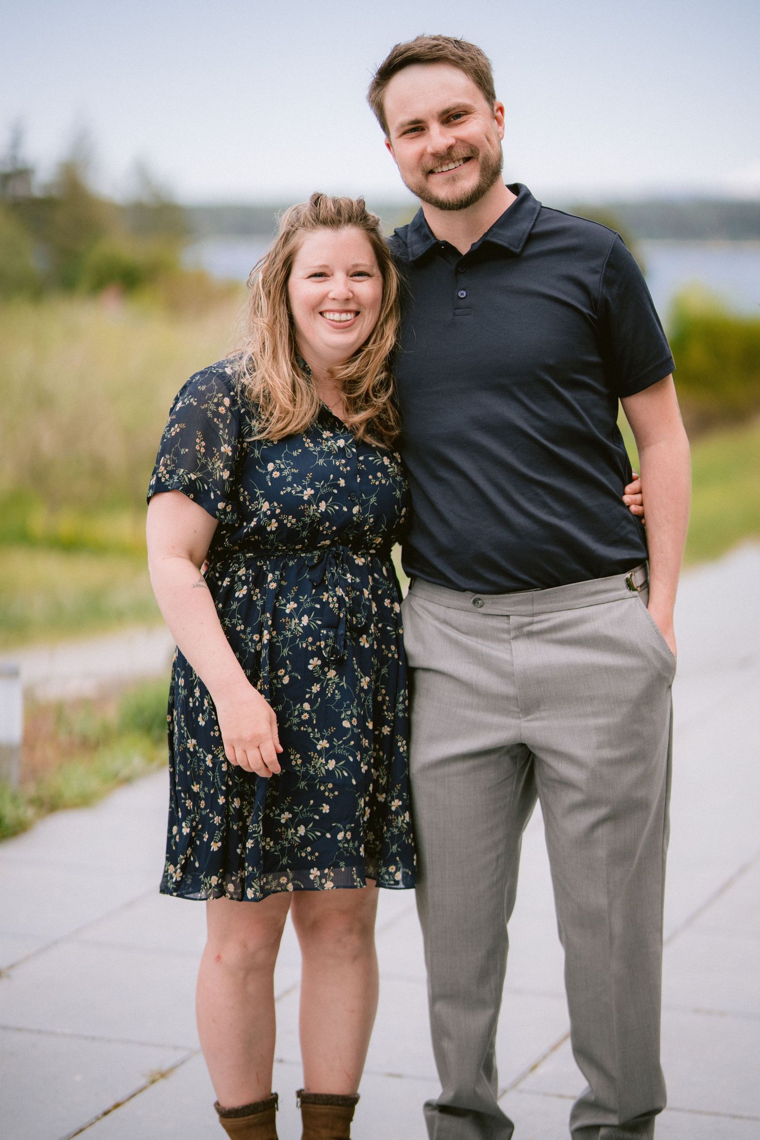 A smiling couple poses together on a paved path with water and greenery in the background.