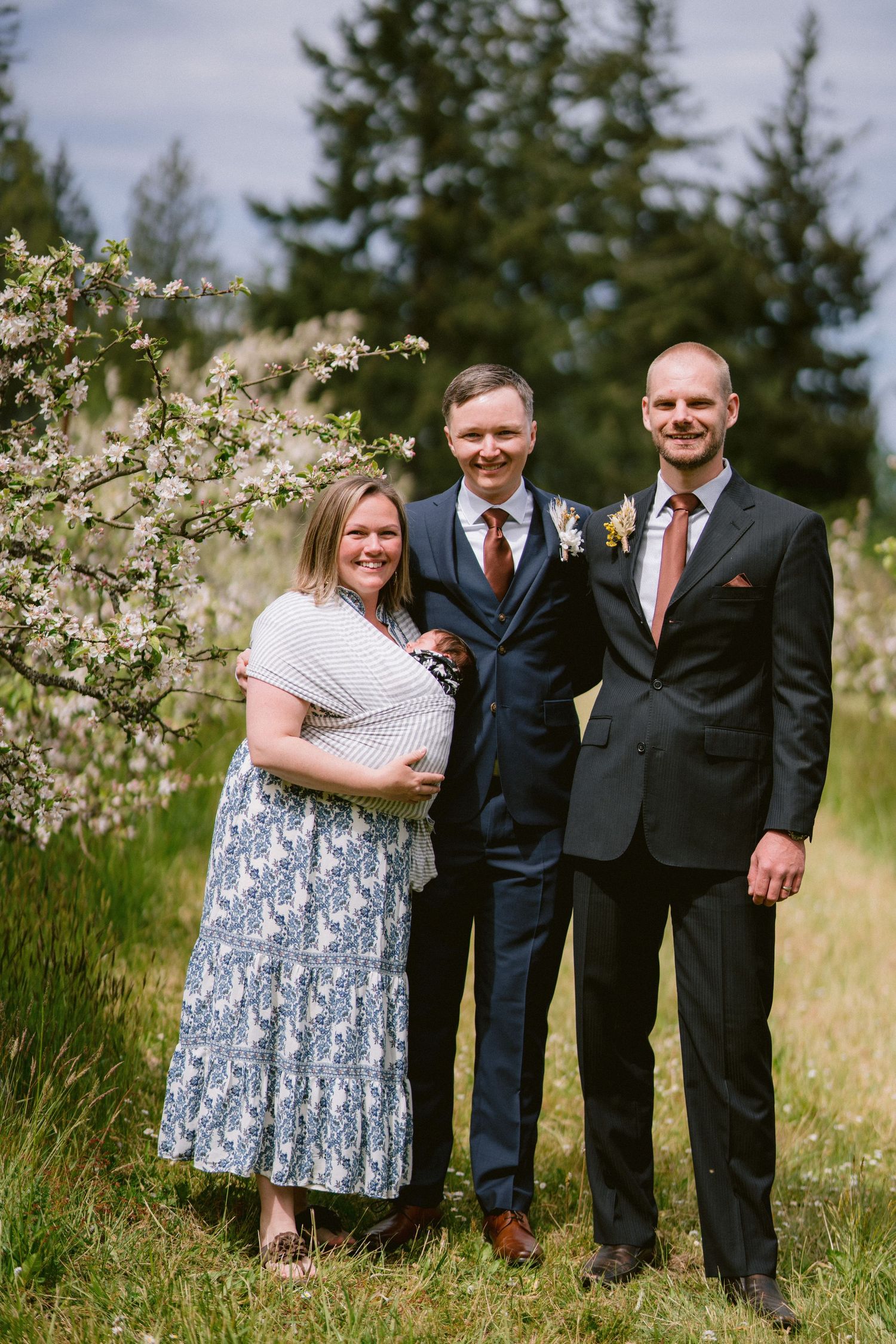 Three people in formal attire pose outdoors near blooming white flowers and evergreen trees on a sunny day.