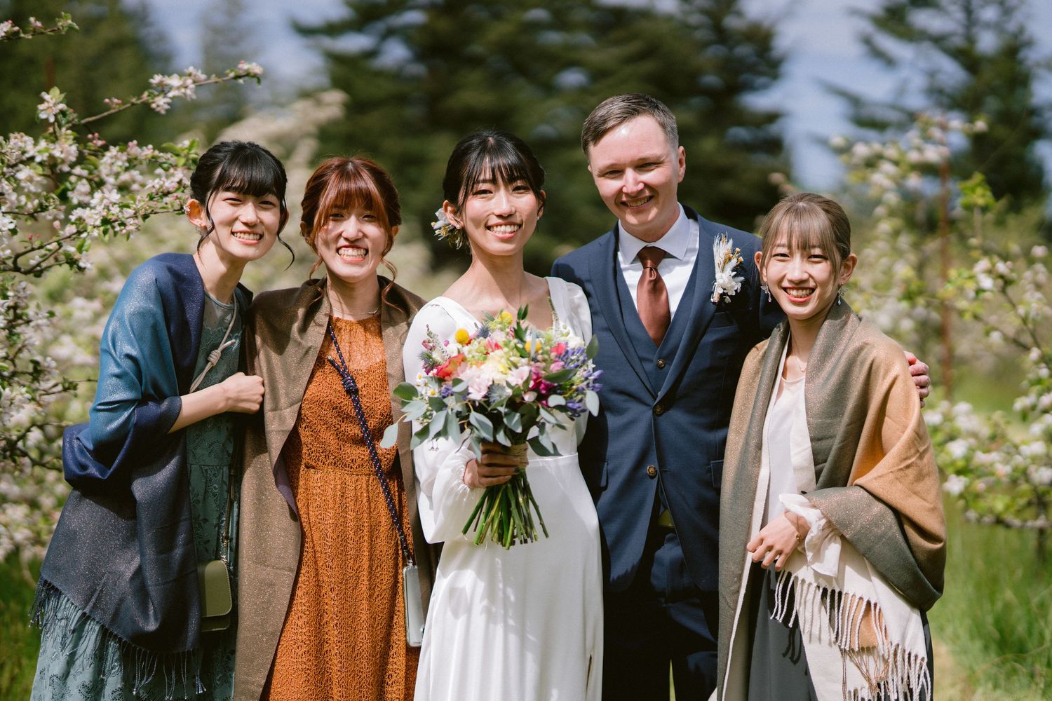 A wedding party poses together in a field of white flowers with evergreen trees in the background.