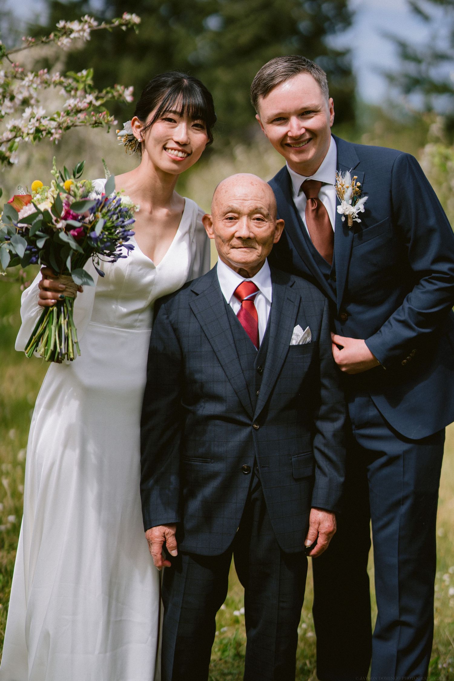 A wedding party poses together outdoors with a colorful bouquet and flowering trees in the background.