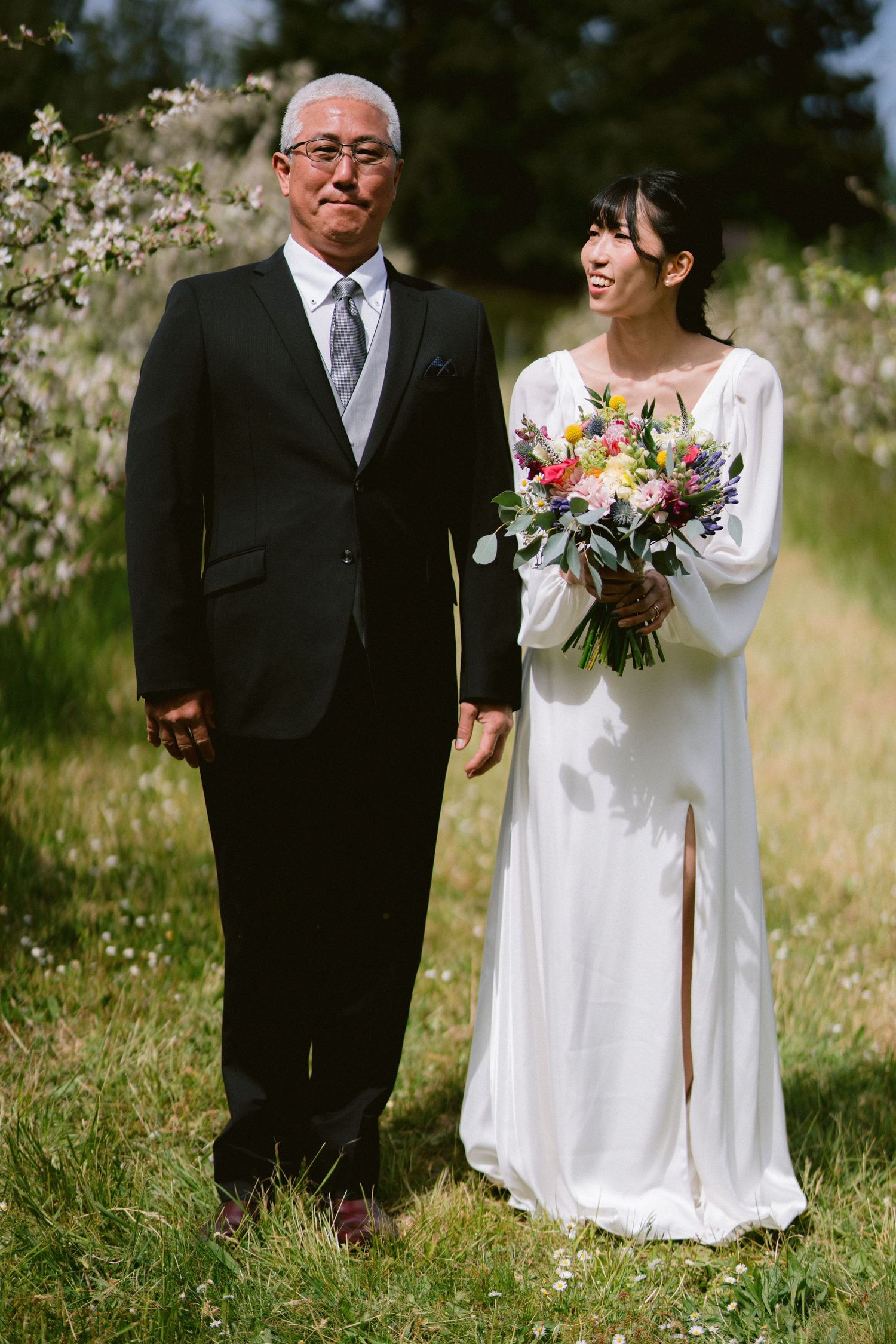 A bride in a white long-sleeved gown holds a vibrant bouquet while standing next to an elegantly dressed companion outdoors.