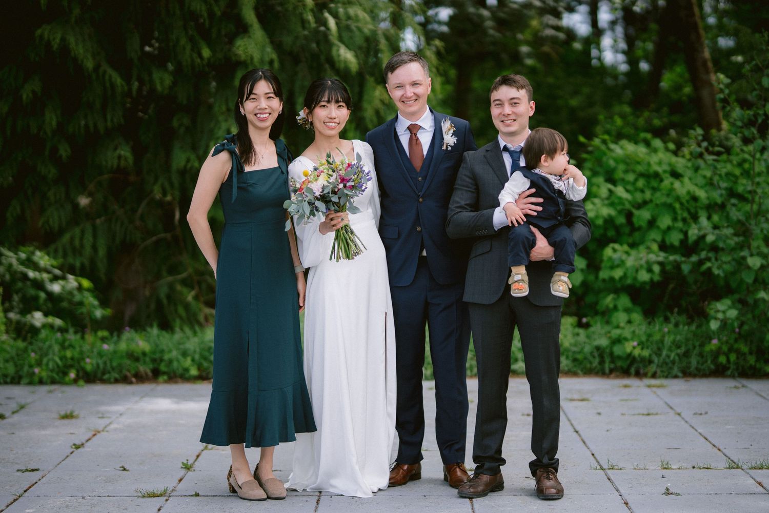 A small wedding party poses together outdoors with greenery in the background.